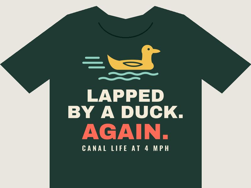 Dark green T-shirt preview with a yellow duck speeding over teal ripples above the words LAPPED BY A DUCK. AGAIN. and CANAL LIFE AT 4 MPH.